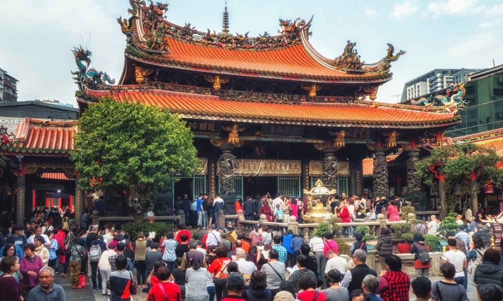 ฝูงชนรวมตัวกันที่วัดอันวิจิตรตระการตาหลังคาสีแดงและแกะสลักมังกร สถานที่สักการะเทพเจ้าต่างประเทศที่มีชื่อเสียง รายล้อมด้วยต้นไม้และอาคารในเมือง.