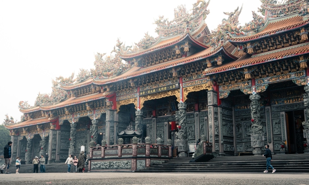 วัดจีนแบบดั้งเดิมที่ประดับประดาอย่างวิจิตร มีลวดลายแกะสลักอย่างประณีต เสาสีแดง และผู้คนเดินอยู่ด้านหน้า—สถานที่สักการะเทพเจ้าต่างประเทศที่มีชื่อเสียง เป็นที่ชื่นชมของผู้มาเยือนที่แสวงหาประสบการณ์ทางจิตวิญญาณ.