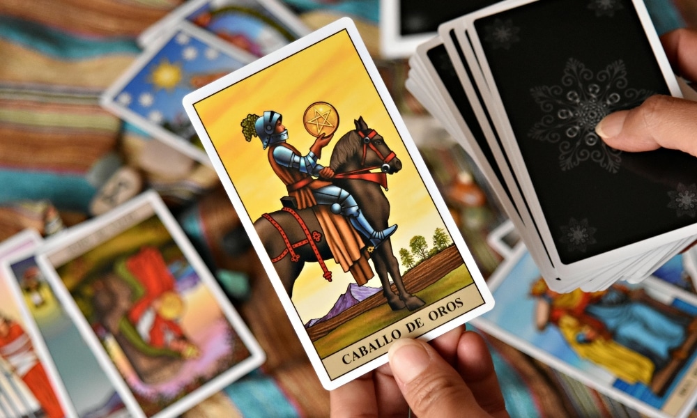มือหนึ่งถือไพ่ทาโรต์ "Caballo de Oros" อยู่เหนือโต๊ะที่มีไพ่ทาโรต์อื่นๆ กระจัดกระจายอยู่รอบๆ บ่งบอกถึงคำทำนายดวงการงาน 12 ราศี เดือนพฤษภาคม.